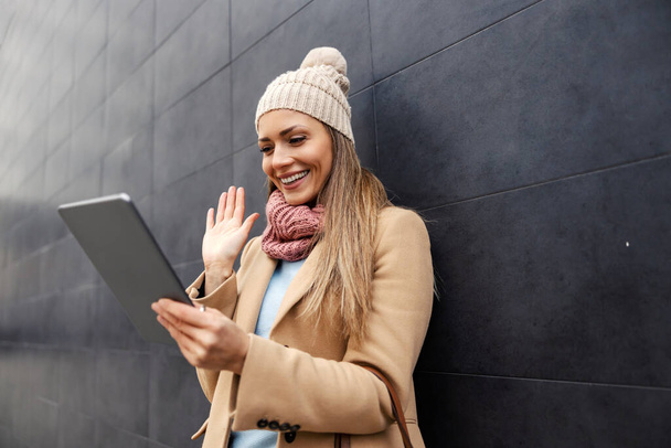 Benefits of the Internet and modern telecommunications. A young happy woman in a warm outfit leaning on the wall outdoors, having a video call on tablet, and waving. - Photo, Image