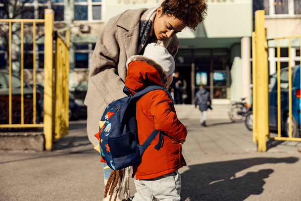 Uma jovem mulher ou uma babá montando o menino para o primeiro dia no jardim de infância enquanto estava em pé em um pátio da escola. - Foto, Imagem