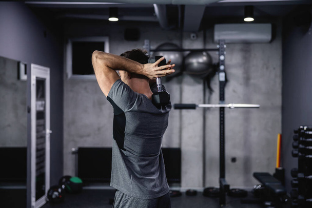 The man keeps good physical shape and fitness. Lifting dumbbells between the shoulder blades in a closed gym with a gray interior. Sports routine and healthy lifestyle - Photo, Image