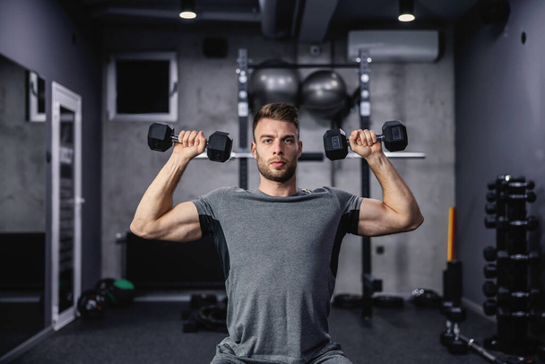 Muscular guy lifting dumbbell while sitting on a bench at the gym. A young athlete using dumbbells during a workout. Strong man under physical exertion pumping up bicep muscle with weight - Фото, изображение