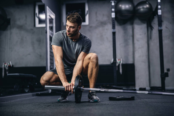Preparing for strong muscle burning training. A young attractive man in gray sportswear sets up barbell weights in the gym. Fit young man looking focused on practice, sports discipline - Photo, Image