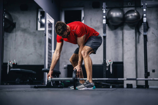 A photo shoot of a handsome man in a red shirt and shorts setting up equipment for training in a modern sports center. Fitness challenge, cross-fit, weightlifting - Photo, Image