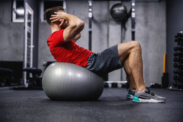 Crunches on Pilates or yoga balls. A muscular and handsome male person in sportswear performs sit-ups on a fitness ball in a modernly equipped dark atmosphere of the sports center - Photo, Image