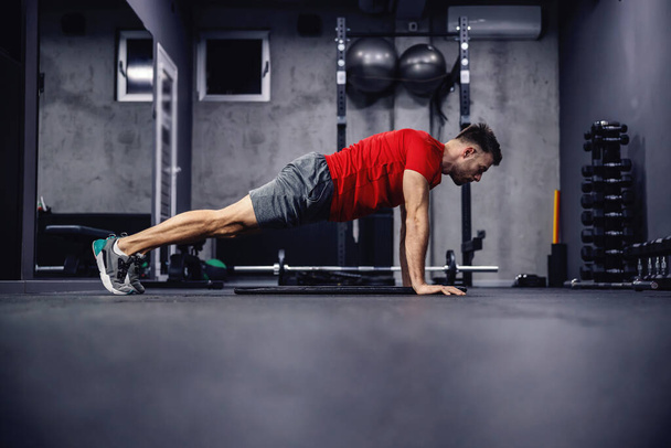 Exercises for arms and shoulders. A man in sportswear with muscles in a modern gym concept does push-ups. He is in a plank position. Empty space for an advertising message - Photo, Image
