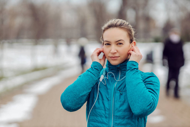 Portrait of sportswoman in warm outfit standing in public park on snowy weather and putting earphones. Winter sport, music, recreation, technology - Photo, Image