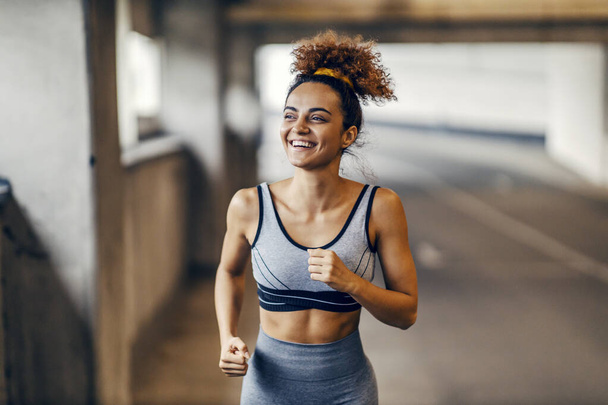Una corredora feliz está corriendo bajo el paso subterráneo con una sonrisa en su cara con el mínimo esfuerzo. Hábitos saludables - cuerpo sano. Un corredor urbano corriendo en la carretera. - Foto, imagen