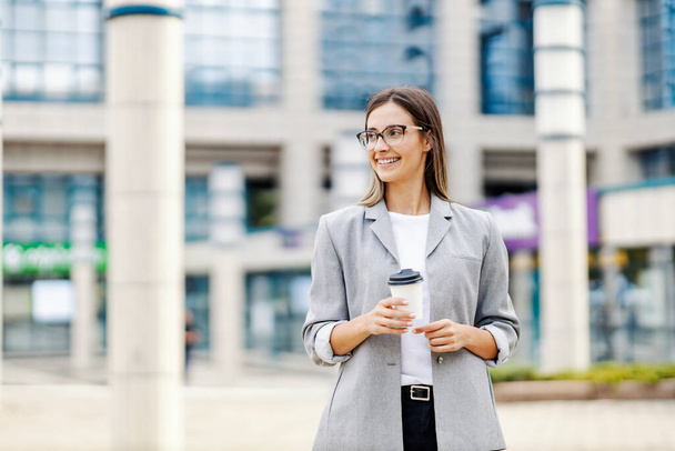 Una mujer de negocios feliz vestida elegantemente, parada en la calle y sosteniendo café para llevar. Una mujer de negocios tomando un café afuera - Foto, Imagen