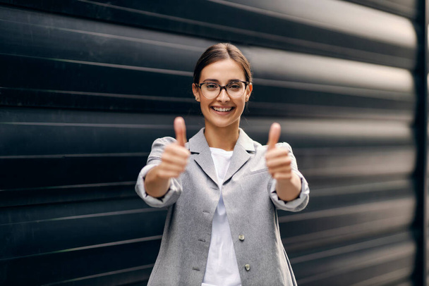 A happy successful businesswoman standing outdoors and showing thumbs up with both hands. The business is going very well - 写真・画像