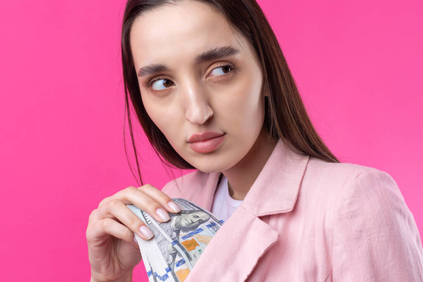 Kobieta w różowej kurtce trzyma banknoty dolarowe, zakłopotana bizneswoman chowa dolary stojące na czerwonym tle. - Zdjęcie, obraz