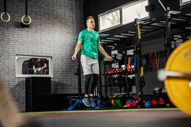 Jumping rope. Athlete man working out on a skipping rope. Beautiful sports male person doing exercises with jumping rope in modern gym space with colorful equipment, worming up for training - Fotó, kép