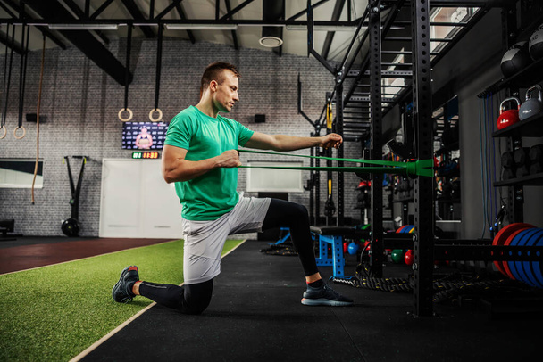 Exercises with straps, flexibility, and body stability. A man in sportswear leaning on one knee with his hand stretches an elastic rubber band and exercises his arms in modern gym space - Photo, Image