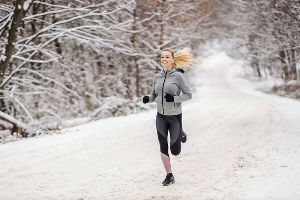 Vrolijke sportvrouw die 's winters op besneeuwd pad door de natuur rent. Cardio-oefeningen, winterfitness, gezonde gewoonten - Foto, afbeelding