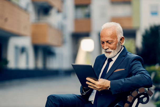 Ein alter Geschäftsmann benutzt in einem Park ein Tablet. Ein glücklicher alter Geschäftsmann sitzt auf der Bank im Park und tippt auf seinem Tablet. - Foto, Bild