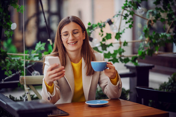 Lächelnder Influencer im gelben Pullover sitzt auf der Terrasse eines Cafés, hält eine Kaffeetasse in der Hand und checkt per Smartphone, was in den sozialen Medien neu ist. - Foto, Bild