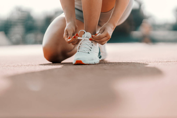 Спортивні жіночі туфлі в тренуваннях. Жінка в спортивному вбранні присідає і пов'язує шнурок. Сірий гардероб і білі кросівки на відкритому тренувальному стадіоні, близький знімок жіночого тіла
 - Фото, зображення