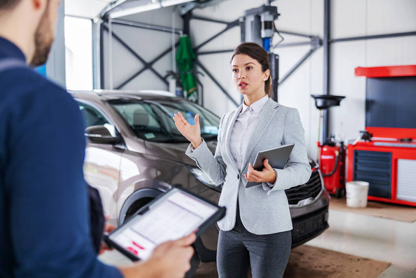Female car seller standing in garage of car salon and talking to a mechanic about repairing the car. - Foto, immagini
