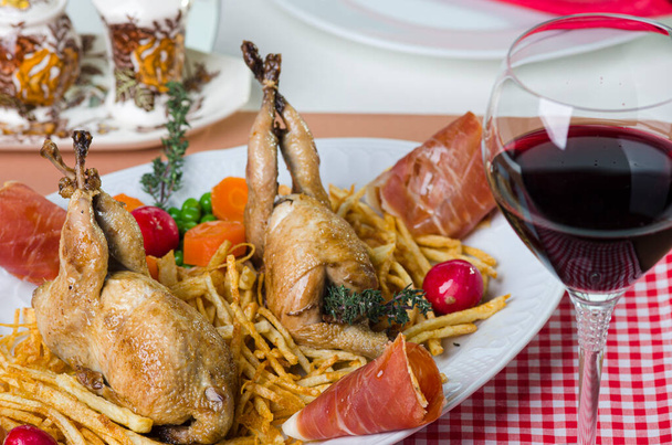 Table with a platter containing 2 quail with fried potatoes, garnished with radishes, carrots and Serrano ham. - Foto, Imagem