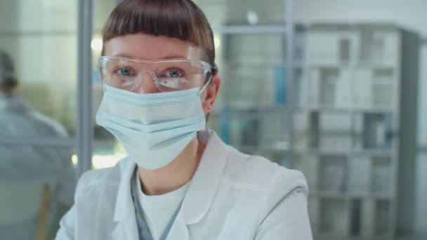 Porträt einer jungen Wissenschaftlerin mit Schutzmaske und Brille, die im Labor sitzt und vor der Kamera posiert - Filmmaterial, Video