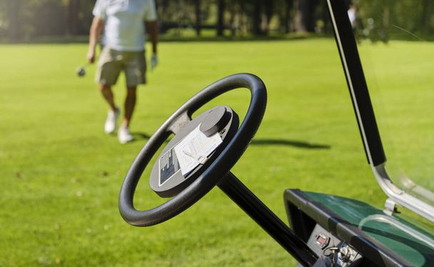 選手はゴルフコースで得点を保持します。スコアは、ゴルフ車のステアリングホイールに置かれます。背景では、手にクラブを持つプレーヤーはゴルフ車を運転する準備ができています. - 写真・画像
