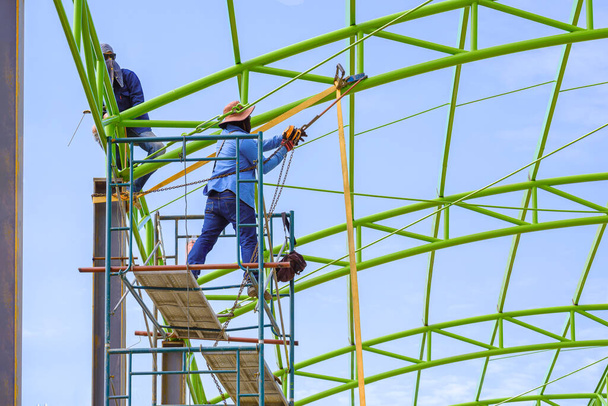 Zwei Bauarbeiter auf einem Gerüst reparieren vor blauem Himmel die grüne Metalldachkonstruktion eines Lagergebäudes  - Foto, Bild