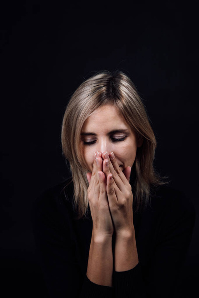 Frightened woman with hands hiding nose and mouth being silent dressed in black blouse on black background. Victim of physical and psychological abuse. Gaslighting. Relative aggression - 写真・画像