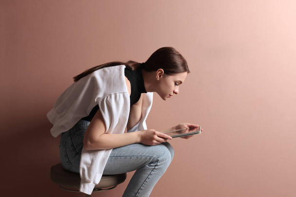 Young woman with bad posture using tablet while sitting on stool against pale pink background - Foto, Bild