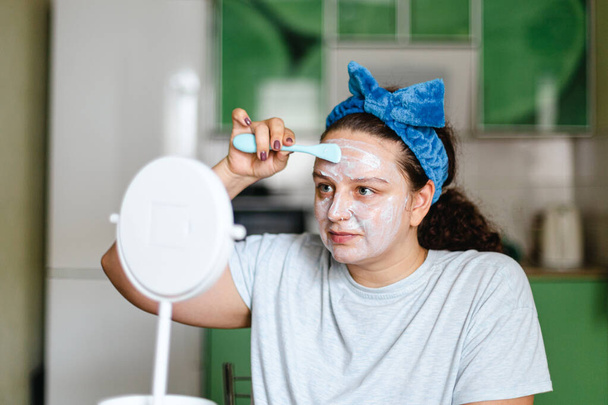 Caucasian young woman putting moisturizing mask on face skin with rubber brush having blue bow on head looking at mirror. Talking care of yourself and skin. Anti age. Home beauty makeup procedures - Foto, immagini