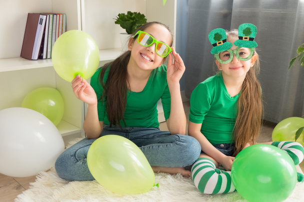 Due ragazze sorelle di 12 e 7 anni in divertenti bicchieri verdi, vestite con magliette verdi, tengono palloncini e si divertono a casa a festeggiare il giorno di San Patrizio. - Foto, immagini