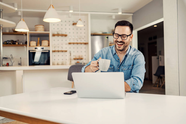 Счастливый человек сидит дома и держит кружку с кофе, улыбаясь в ноутбук. - Фото, изображение