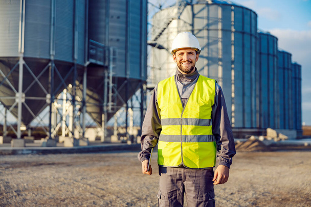 Portret pracownika przemysłu trzymającego tablet przed silosami pełnymi ziarna i uśmiechającego się do kamery. - Zdjęcie, obraz