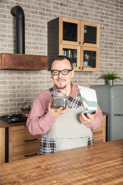 Funny handsome young smiling man wearing pinafore with towel smell jar with coffee beans on the kitchen - Photo, Image