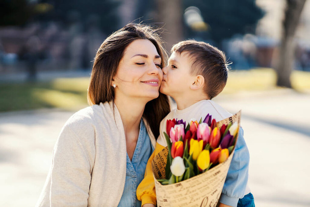 Маленький мальчик дарит букет тюльпанов своей матери и целует ее в день матери.. - Фото, изображение