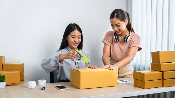 Две молодые предпринимательницы упаковывают товар в коробку, курьеры подтверждают заказ и перепроверяют адрес клиента на смартфоне для подготовки отправки домой - Фото, изображение