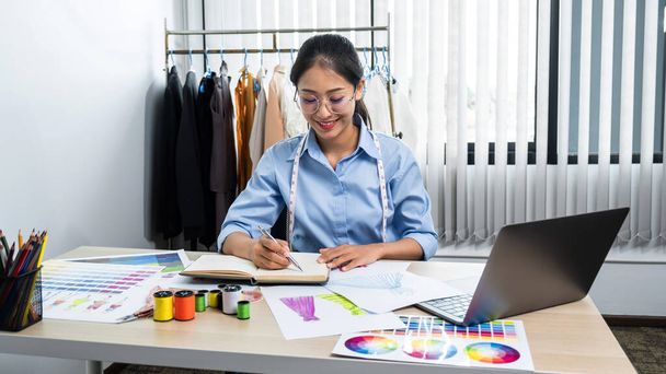 Asian woman fashion designer wearing glasses to drawing clothes sketch and working with fabric while sitting in her workshop - Photo, Image