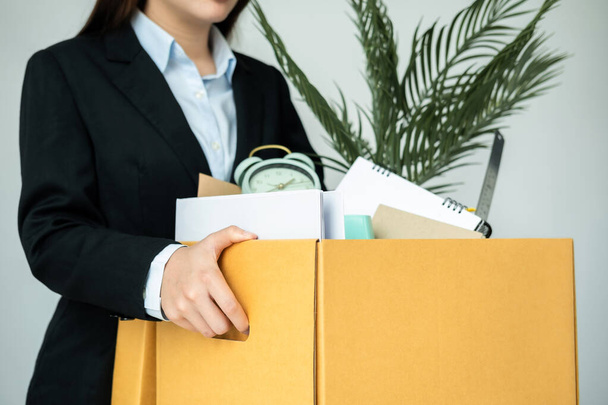 Takım elbiseli iş kadını çalışanı işten istifa ederken özel eşyalarını kahverengi karton kutuya koyar ve ofisten çıkıp gider. - Fotoğraf, Görsel