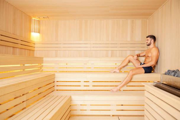 A man in the spa staying healthy. A handsome muscular man in swimwear sitting in a sauna and relaxing. - Foto, Imagem