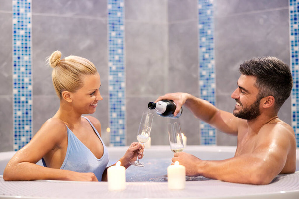 Un couple romantique boit du champagne dans le bain à remous du centre spa. Un mari heureux verse du champagne dans le verre de sa femme tout en étant assis dans le bain à remous au centre de bien-être et de spa avec elle. - Photo, image