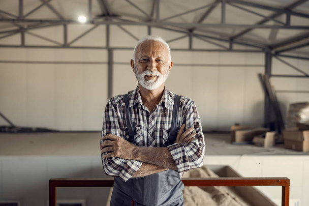 Operai anziani di successo. Un orgoglioso e sorridente operaio anziano in uniforme da lavoro è in piedi in fabbrica con le braccia incrociate.. - Foto, immagini