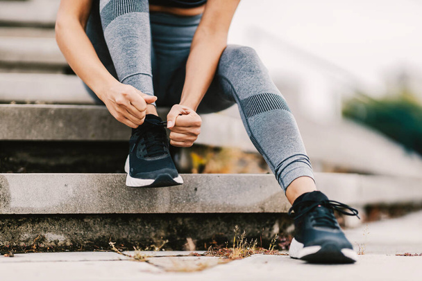 Acercamiento de la corredora sentada en las escaleras al aire libre y atando un cordón de zapato en su zapatilla. Un corredor preparándose para correr - Foto, imagen