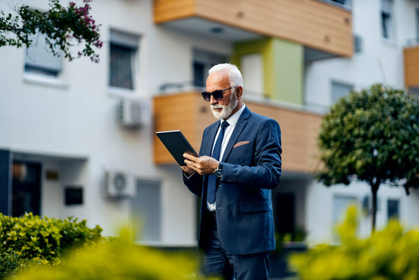 An old businessman in rush using a tablet while walking. A busy serious senior businessman walks in a park and gets work done on a tablet. - Foto, immagini