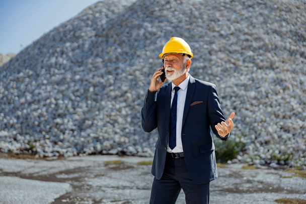 Construction site works and business. A senior businessman with a helmet on his head stands at the construction site and has a phone conversation with the constructor. - Photo, Image