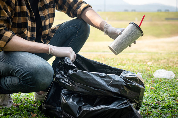 Homem voluntário em luvas sentado para pegar xícara de café em saco preto de plástico para limpar o parque durante a atividade ambiental para coletar lixo - Foto, Imagem