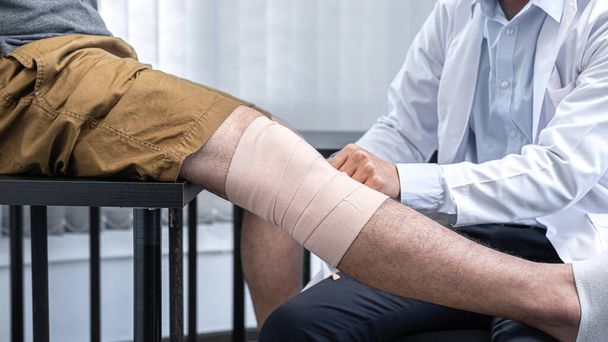 Junger asiatischer Physiotherapeut bandagiert Knie eines Patienten mit Verletzungen während der Heilbehandlung und gibt Rehabilitationstherapien in der Klinik - Foto, Bild