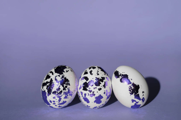 Tres huevos blancos de Pascua decorados con lámina de color azul-púrpura sobre un fondo de color Very Peri. Huevos pintados con estilo de moda. - Foto, imagen