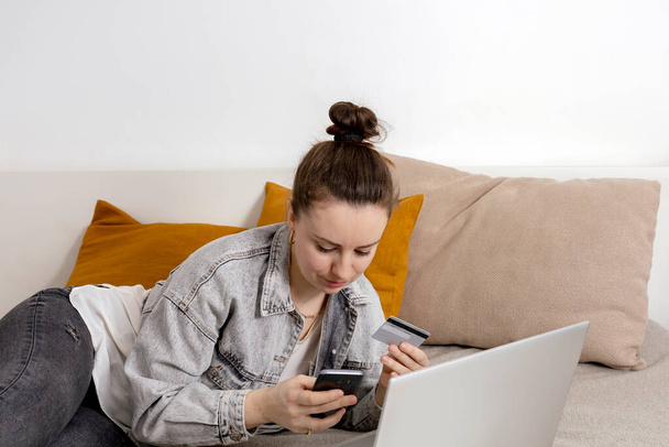 Jeune femme brune souriant tout en surfant sur le web sur son ordinateur portable et tenant la carte de crédit dans sa main en essayant d'acheter quelque chose en ligne. Achats en ligne faciles à la maison, e-commerce. Intérieur confortable. - Photo, image
