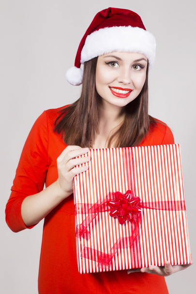 brünette Frau in rotem Kleid und Weihnachtsmannmütze mit Geschenk und Blick in die Kamera - Foto, Bild