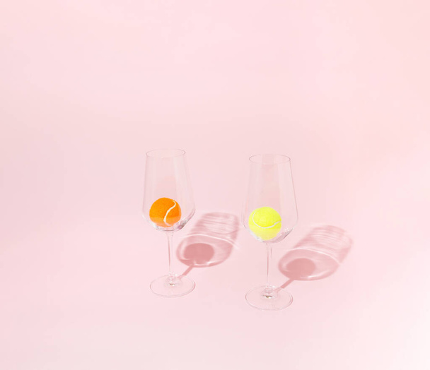 A yellow and an orange tennis ball in two transparent wine glasses on a pink background. Minimal concept. - Fotó, kép
