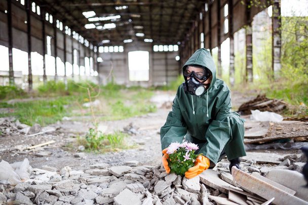 Un hombre con impermeable y máscara de gas recoge una flor de una tierra quemada y tóxica. Concepto de contaminación atmosférica. Catástrofe ecológica. - Foto, imagen