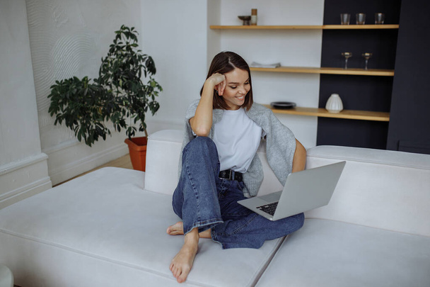 A young brunette girl in a white t-shirt and blue jeans sits at home on a white sofa and uses a laptop - Foto, Imagem