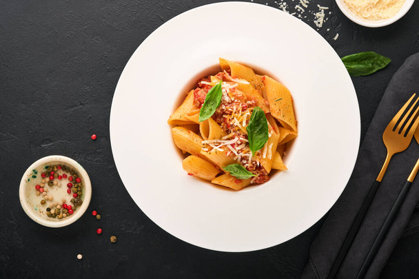 Bolognese Penne Pasta. Tasty appetizing classic italian penne pasta with parmesan, basil and Bolognese sauce in white plate on plate on black dark background.Traditional Italian cuisine. Top view. - Zdjęcie, obraz